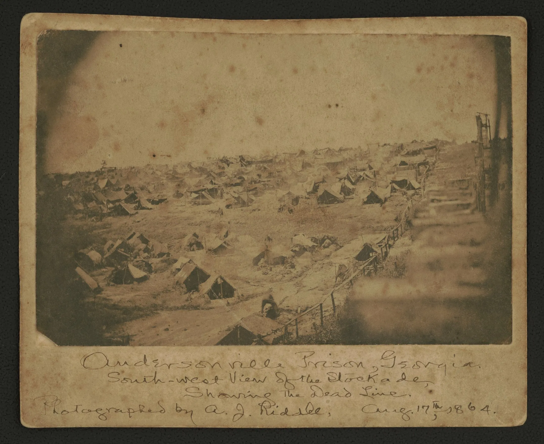A crowded prison stockade seen from a corner, tents and huts packed inside a high timber wall, with a low rail fence running along the inside of the wall.