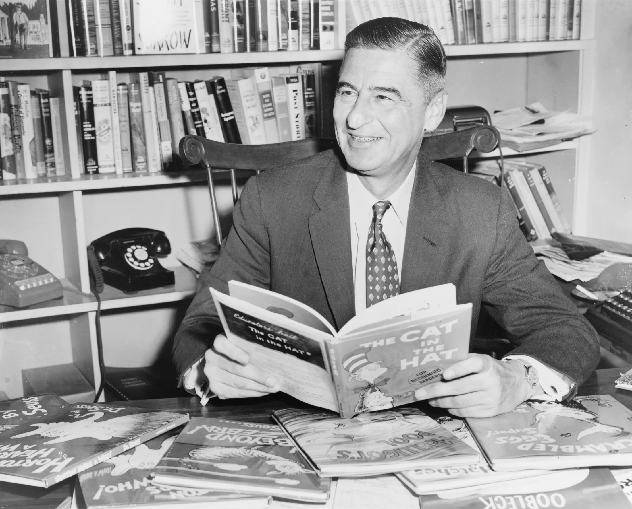 A man in a suit and bow tie sits at a desk piled with picture books, smiling at the camera.