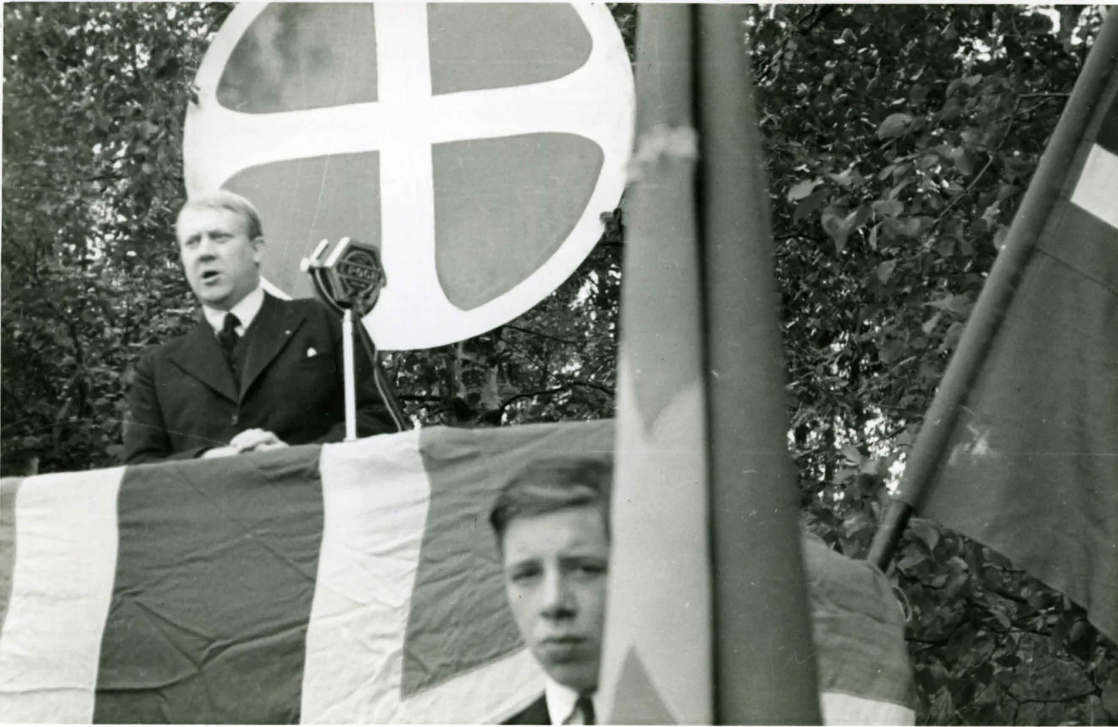 A man in a suit speaks from a lectern draped with a flag, in front of a crowd, with banners behind him.